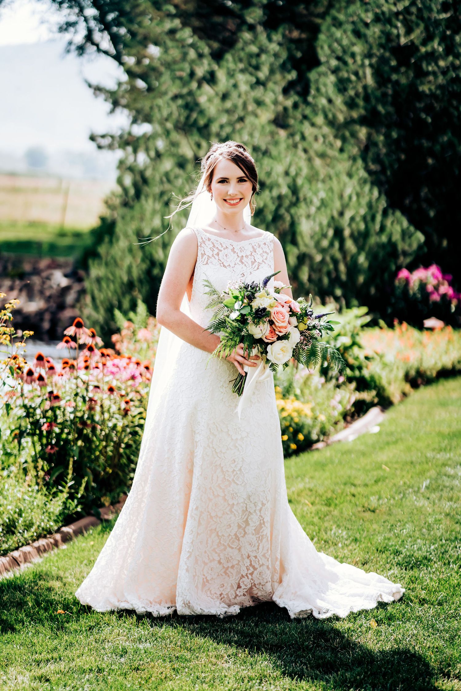 Bride in lace wedding gown holding peach and pink bouquet poses in garden with blooming flowers.