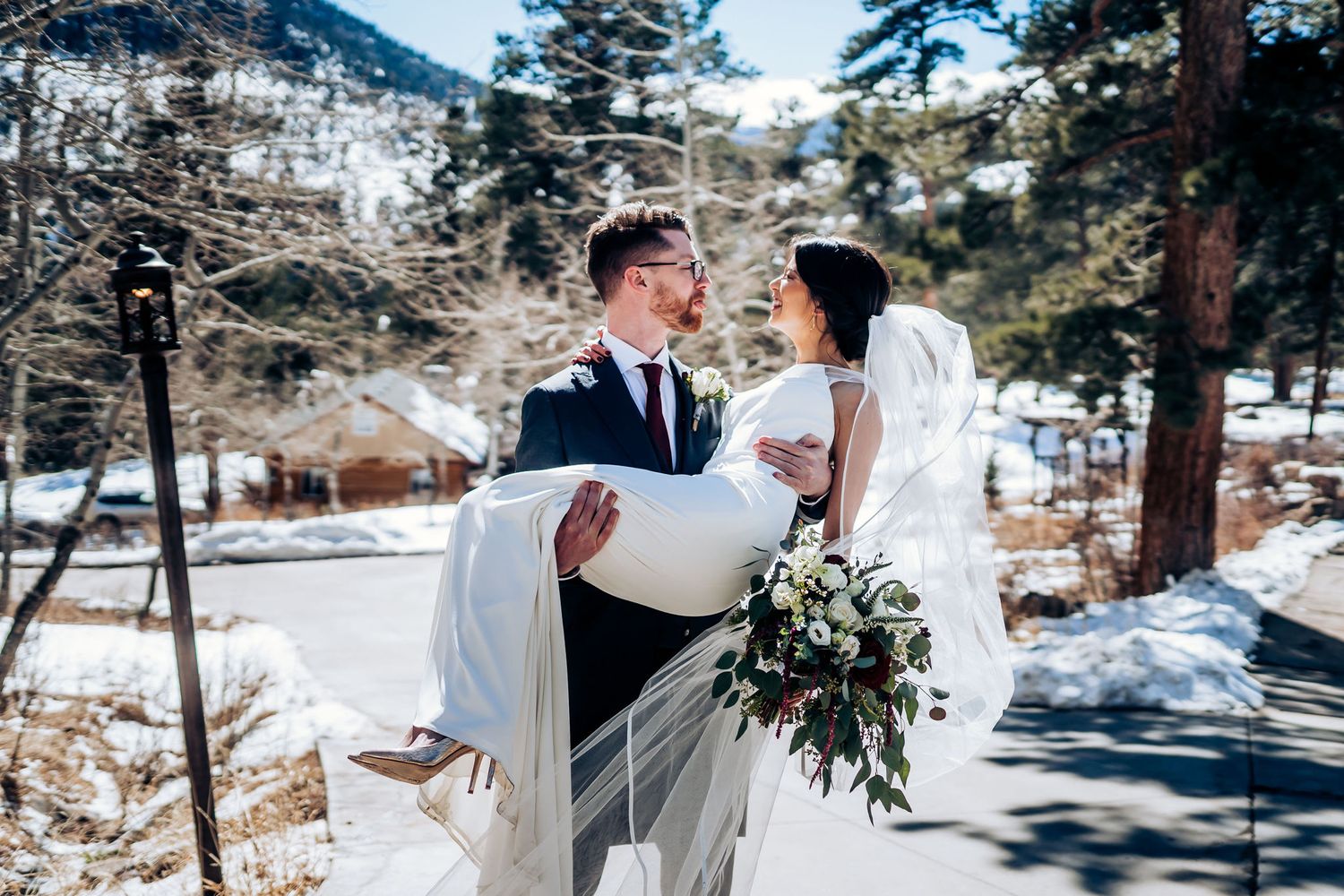 A romantic wedding carry pose in a snowy mountain setting with evergreen trees.