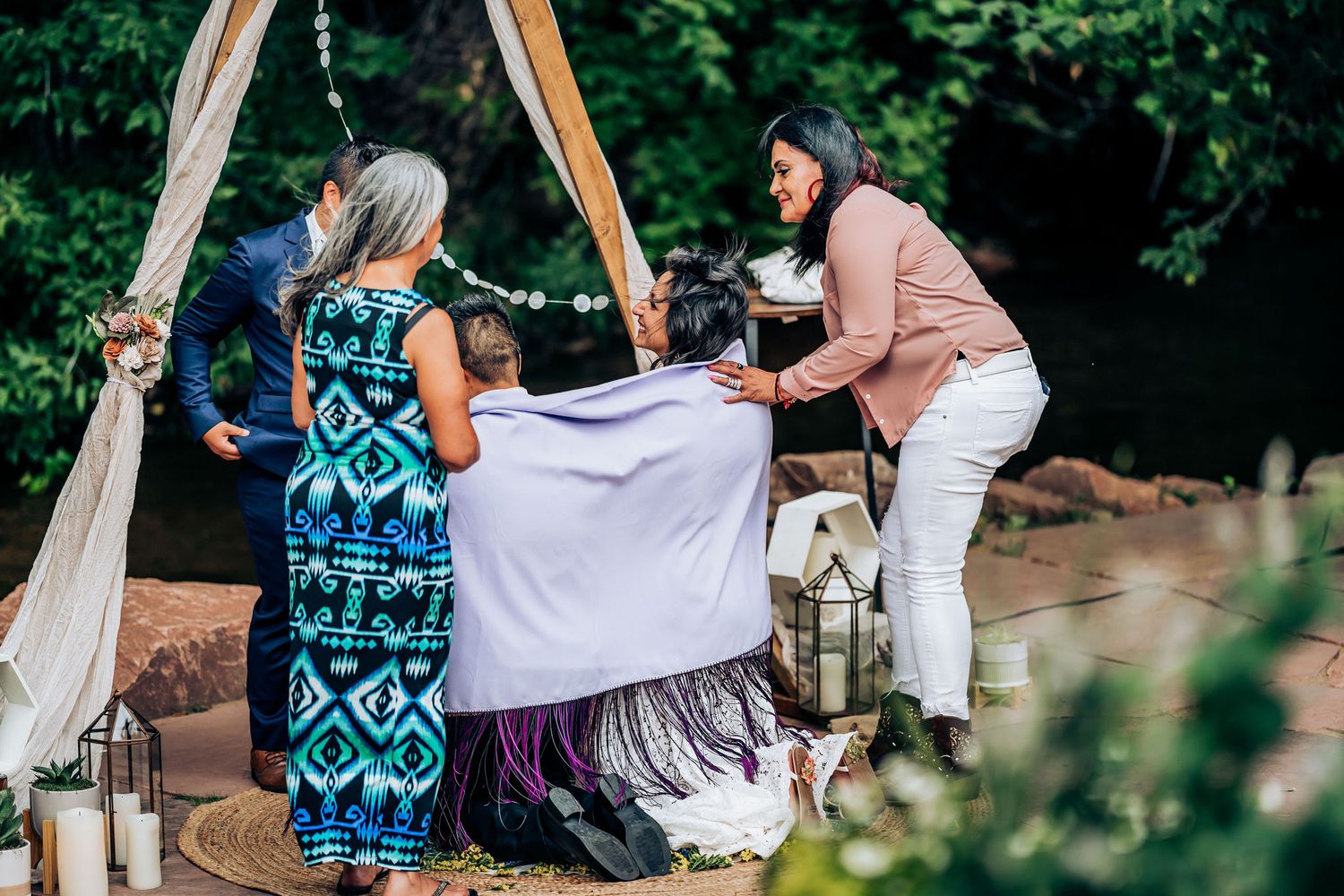 Guests gather around a ceremonial setup with white draping and wooden arch during an outdoor celebration.