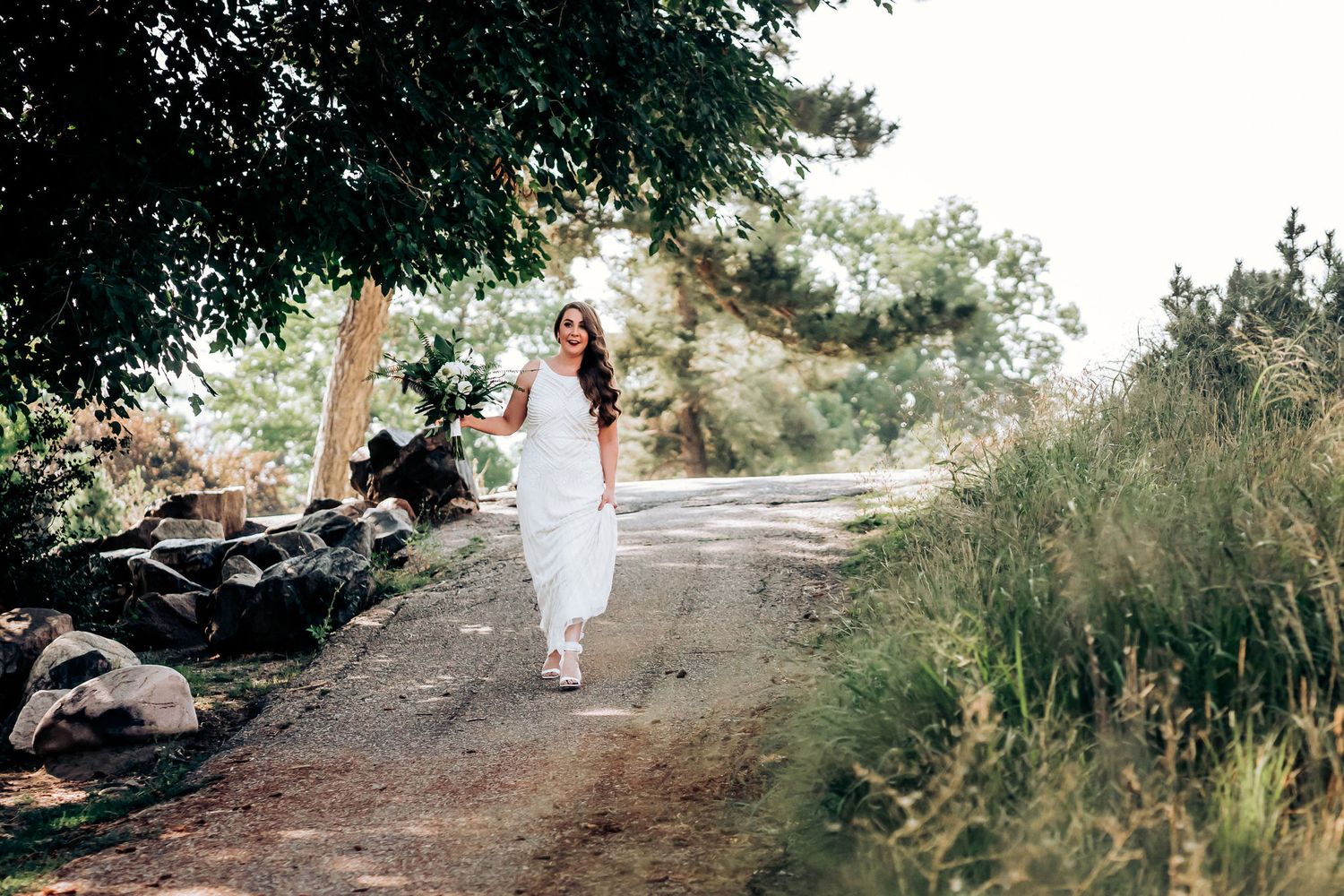 Person in flowing white dress walking along a sunlit outdoor path.