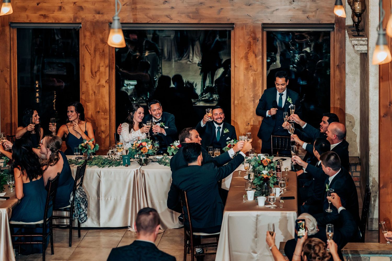 Wedding reception guests applaud during speeches in a rustic wooden venue with elegant table settings.