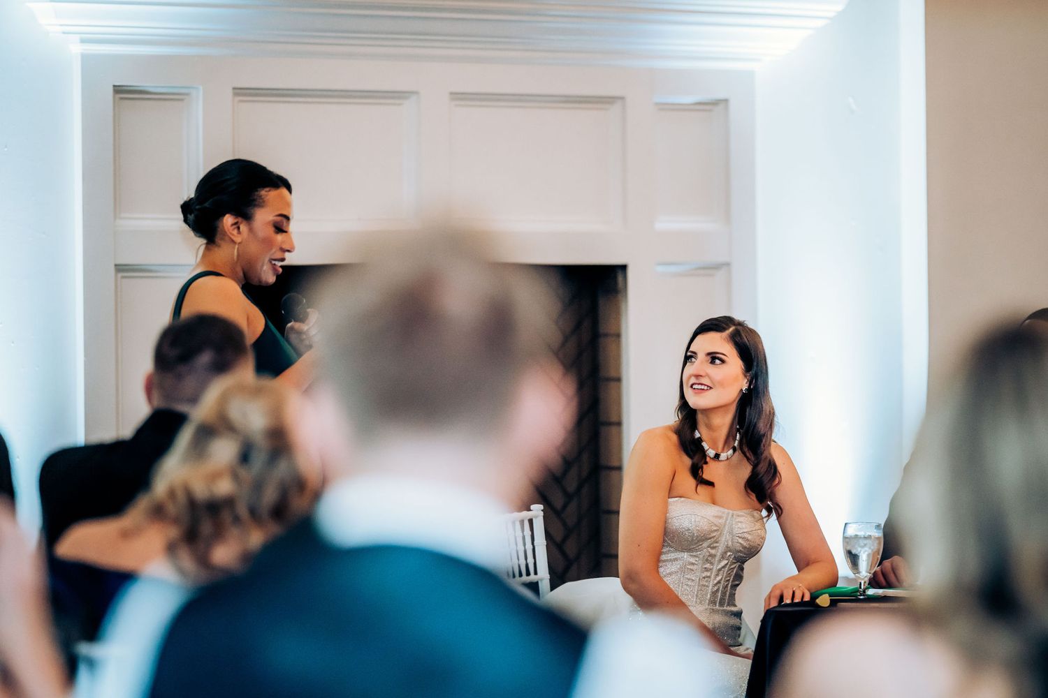 Wedding reception speech in progress at elegant white venue with fireplace backdrop.