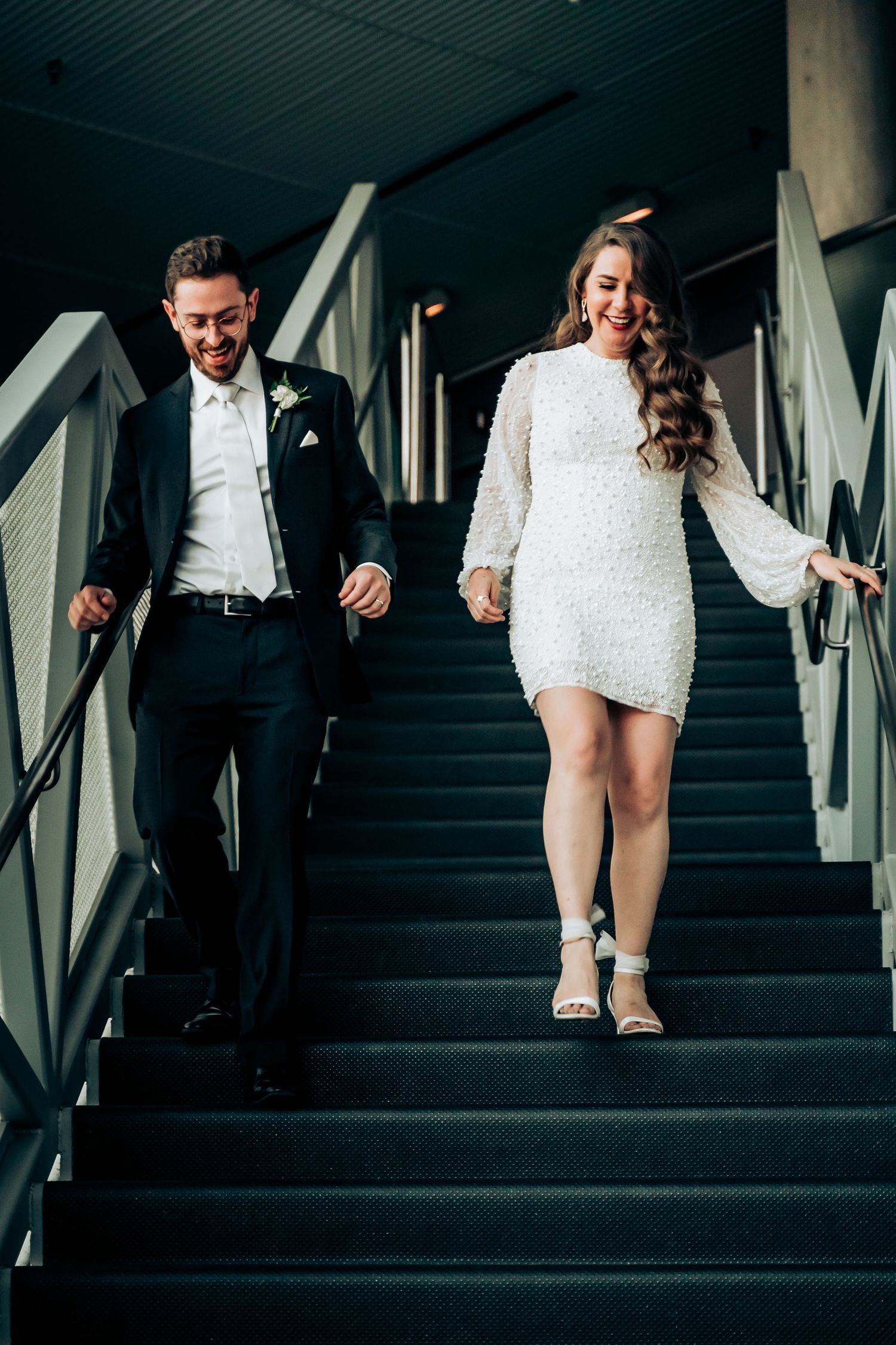 Newlyweds descending modern stairs together, bride in short white dress and groom in dark suit.