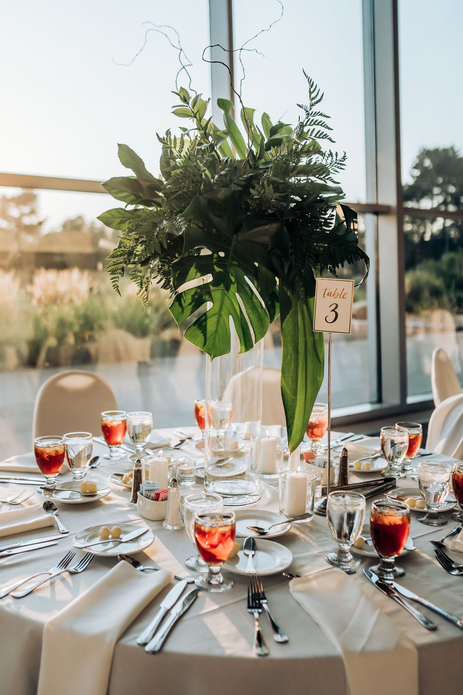 Elegant wedding reception table setting with tall tropical greenery centerpiece and red cocktails.