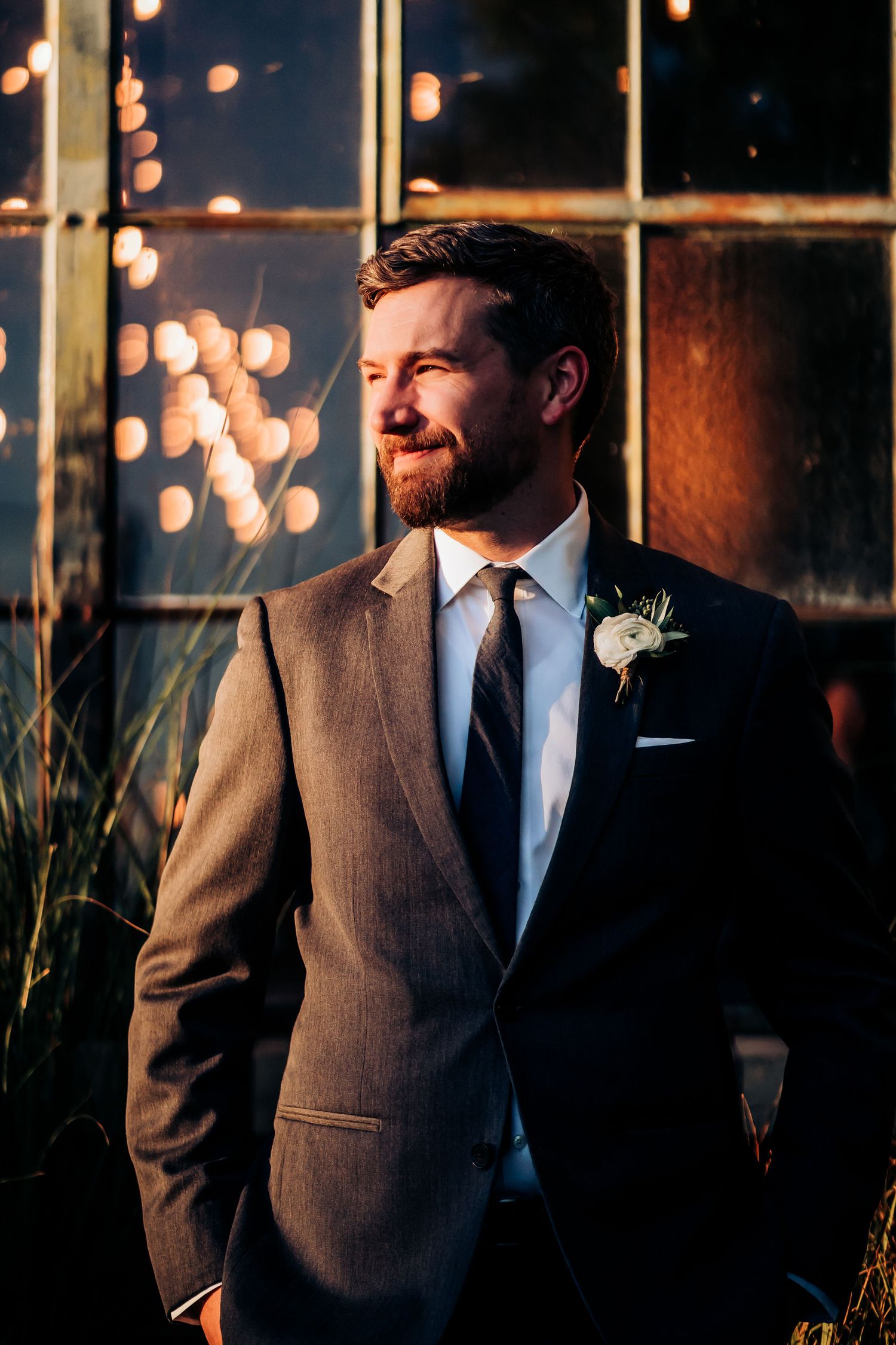 Groom in gray suit posing by window with twinkle lights in background.