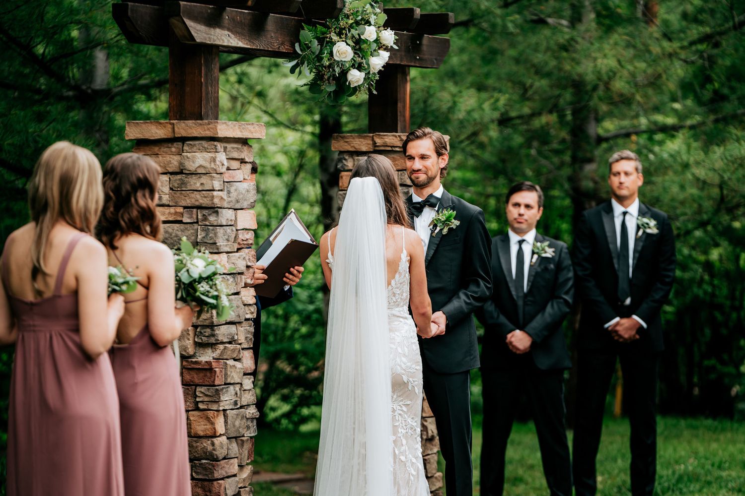 A wedding ceremony under a stone archway with bridesmaids in pink dresses watching the exchange of vows.