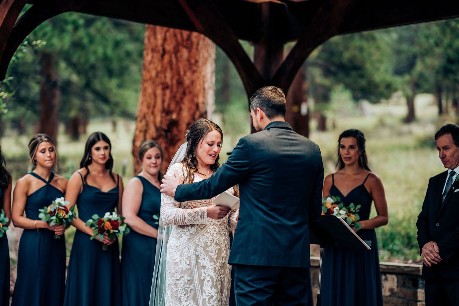 An intimate wedding ceremony takes place under a wooden pergola with bridesmaids in navy dresses.