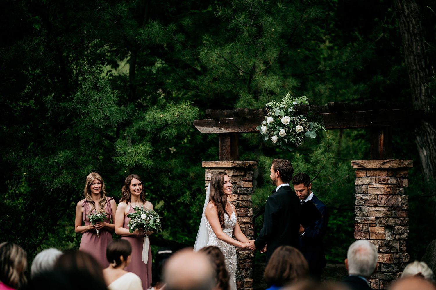 An outdoor wedding ceremony takes place between stone pillars adorned with white floral arrangements.