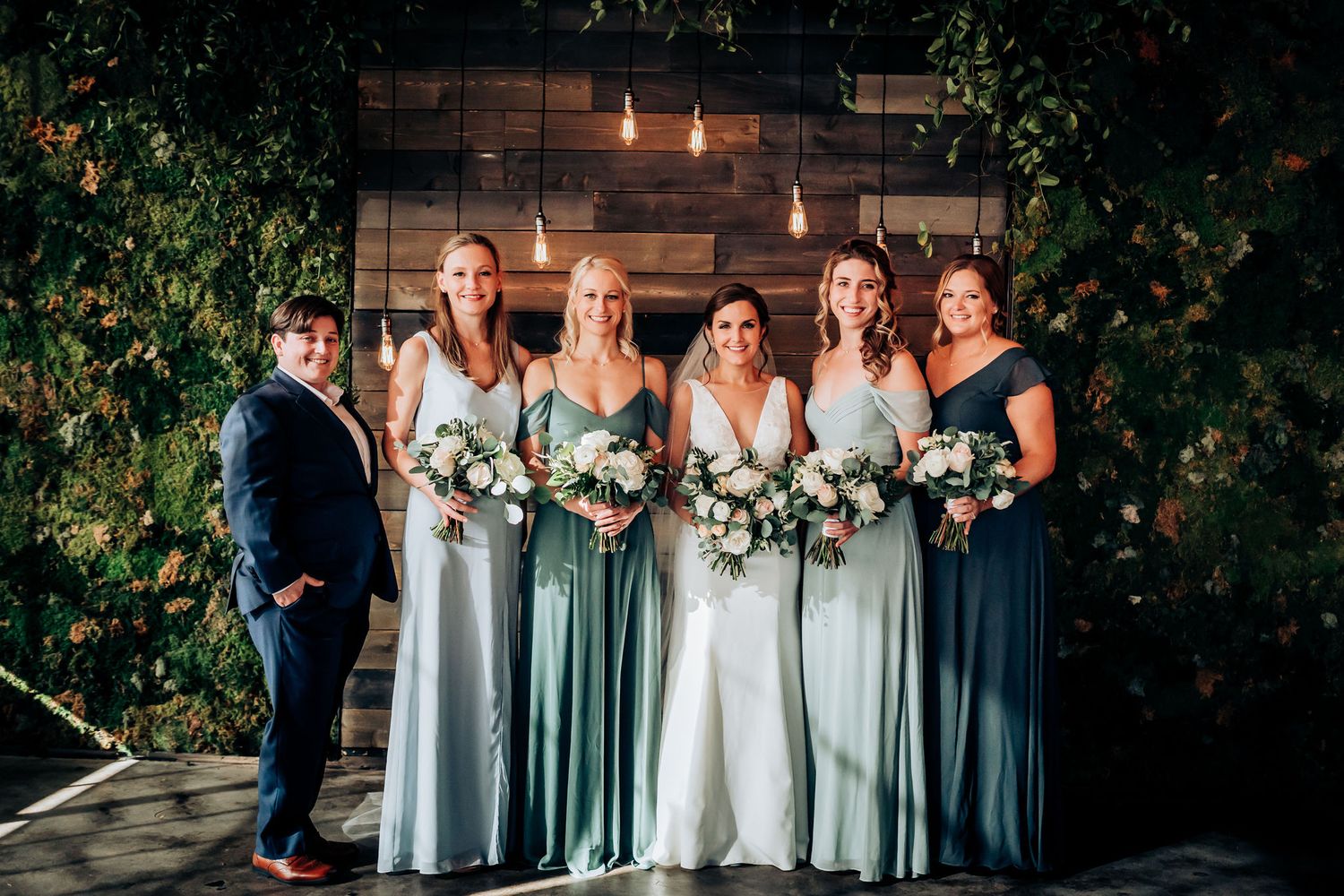 Wedding party in varying shades of blue dresses pose against wooden wall with string lights.