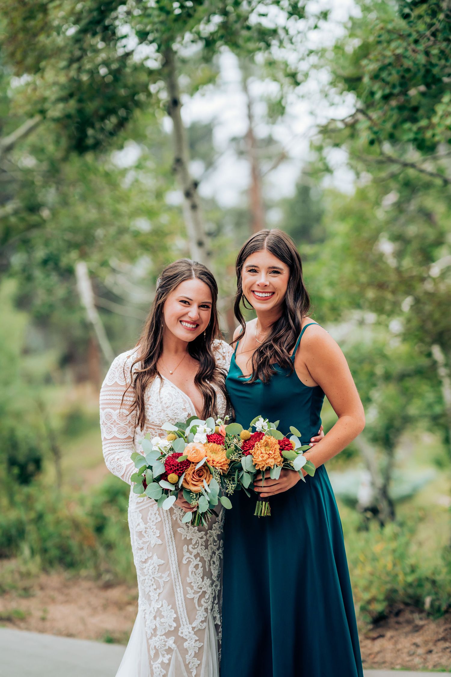 Two wedding attendants in contrasting white and teal dresses hold vibrant floral bouquets outdoors.