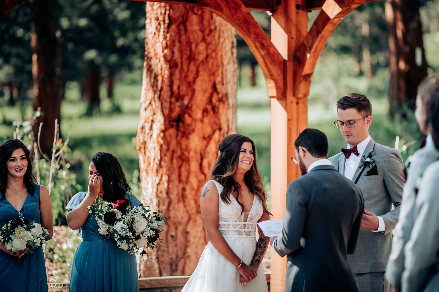 Wedding ceremony takes place beneath wooden archway surrounded by tall pine trees in scenic outdoor setting.