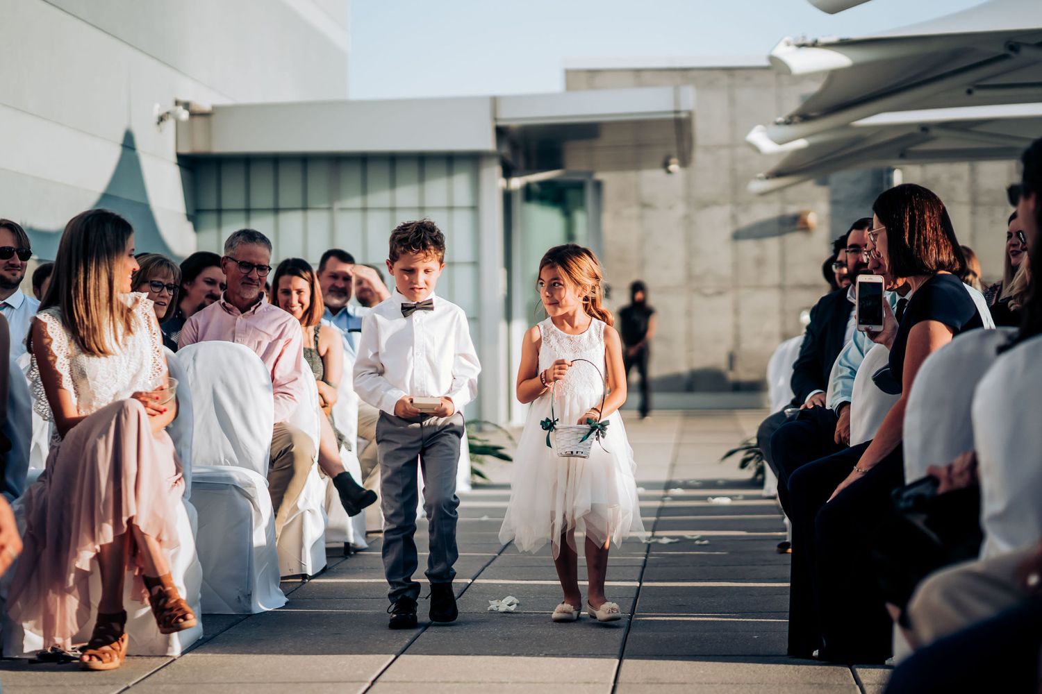 Young attendants walk down an outdoor wedding aisle while guests in white and black attire look on.