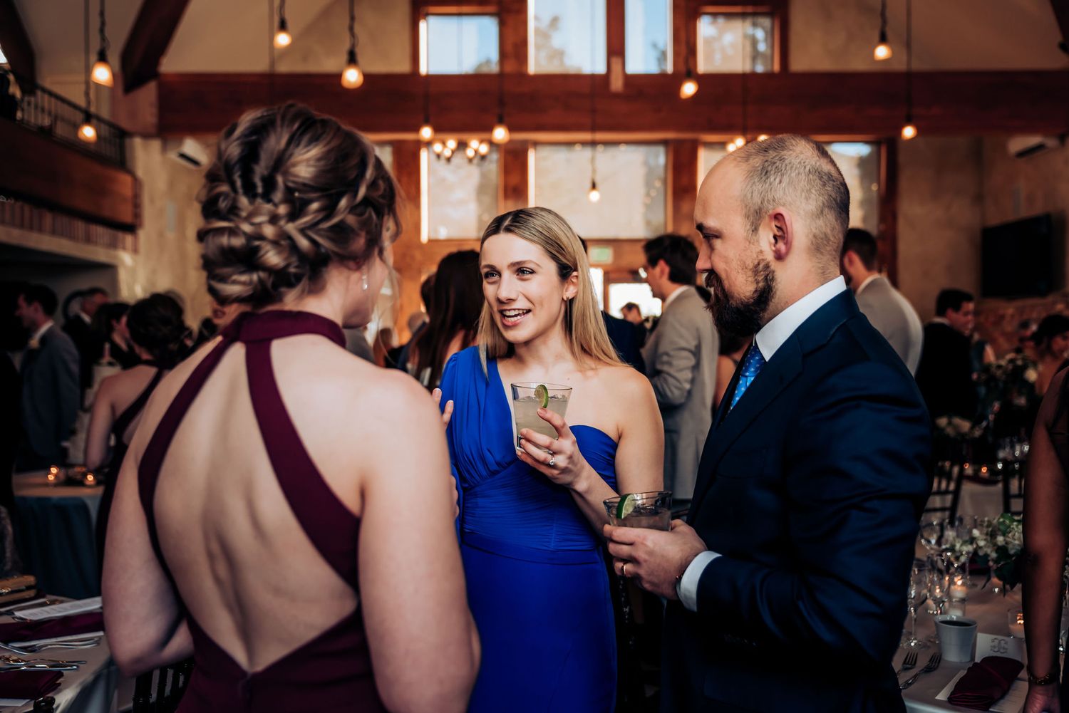 Wedding guests in elegant attire mingle during reception at rustic venue with warm lighting.