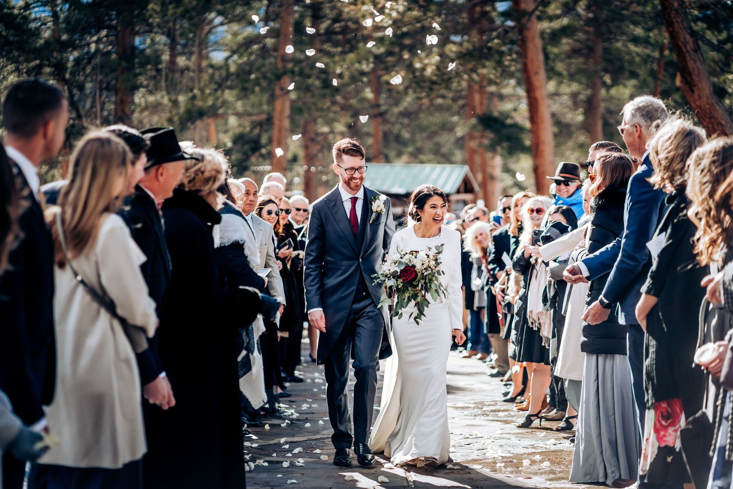 Couple walks down forest path while wedding guests capture moment on phones.