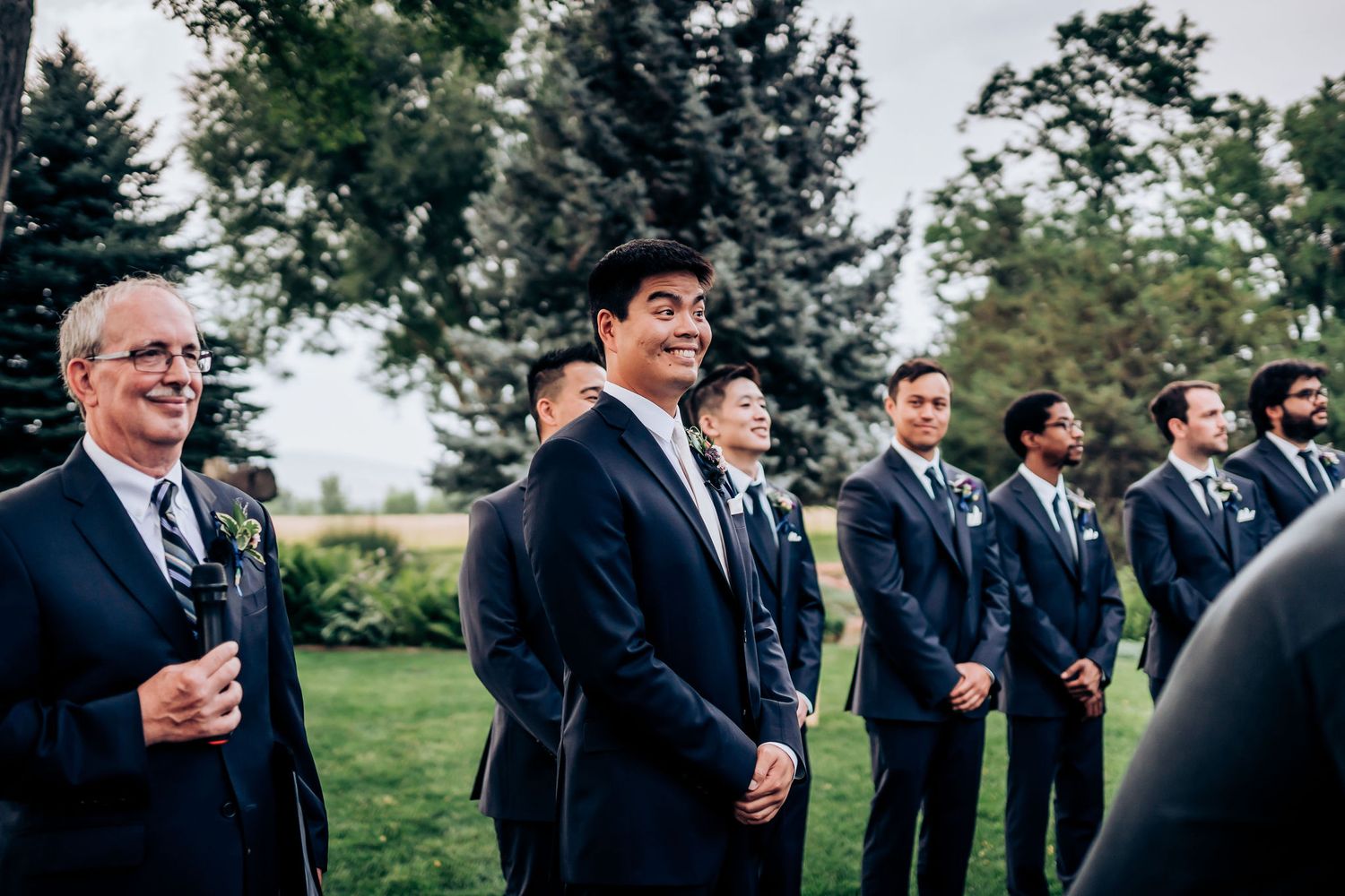 Groomsmen and guests stand outdoors on a grassy lawn during a wedding ceremony with evergreen trees in background.