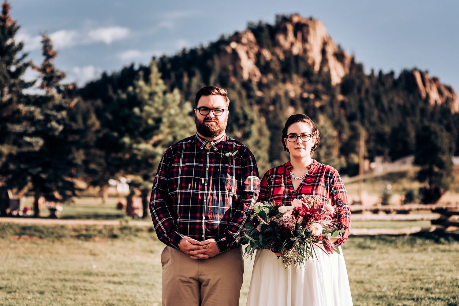 Couple in plaid shirts pose before mountain landscape with bouquet.