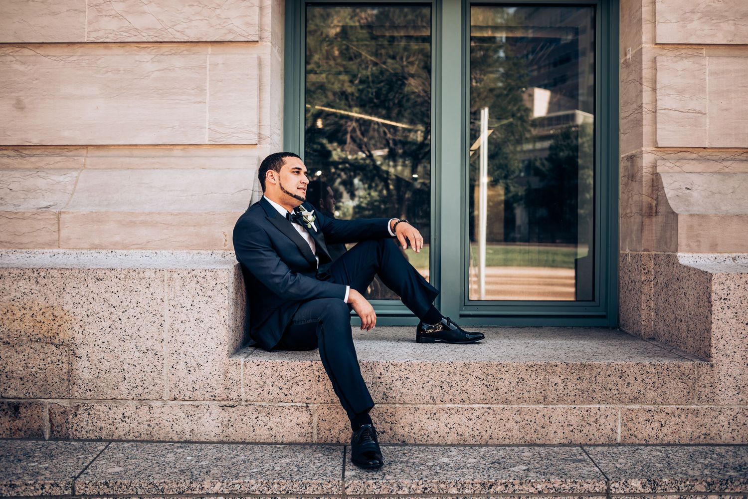 Groom in navy suit sits casually against stone building window ledge.