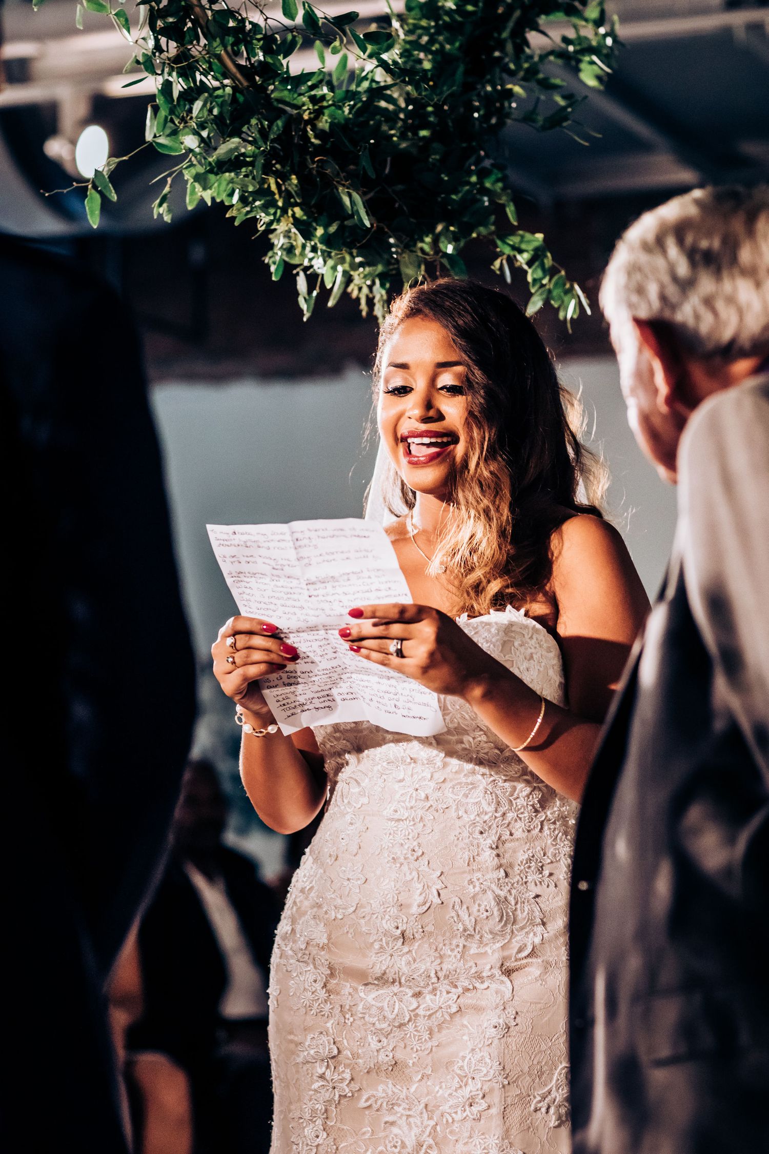 A bride laughs joyfully while reading her wedding vows beneath hanging greenery.