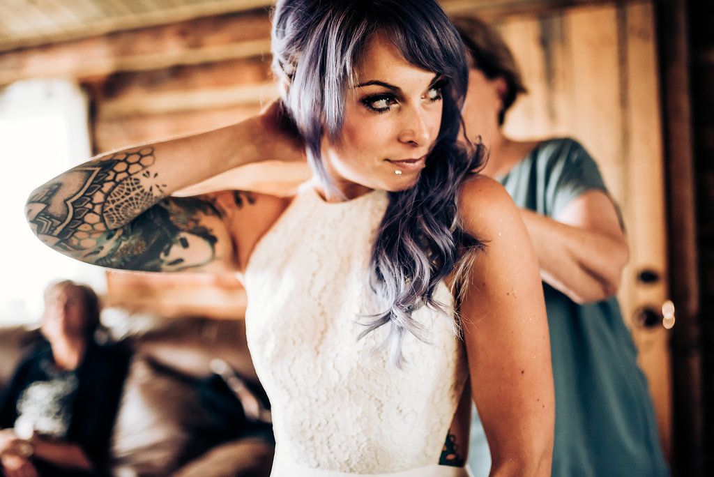 Woman in white dress with visible arm tattoos getting ready in rustic cabin.