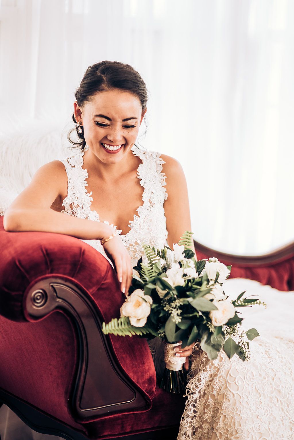 Joyful bride laughs while relaxing on red velvet couch with white bouquet.