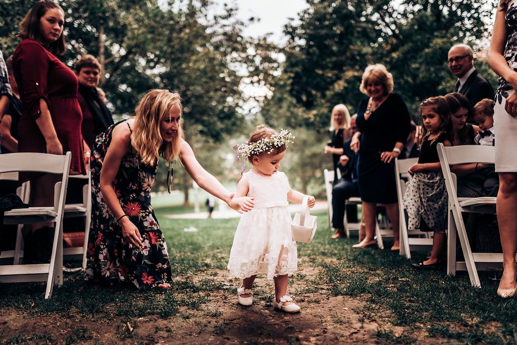 Small flower girl in white dress pauses along wedding aisle while guests look on.