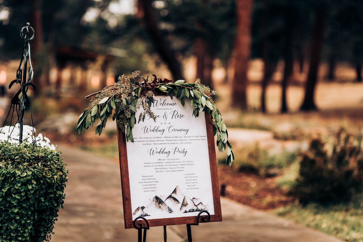 Elegant wedding program sign decorated with greenery stands on easel outdoors.