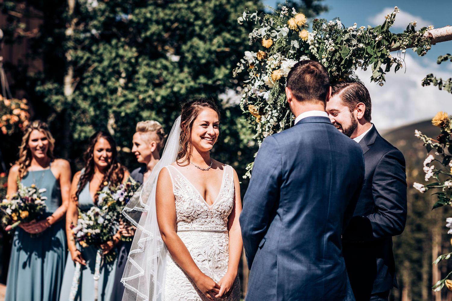 Outdoor wedding ceremony with bridal party in blue dresses standing under floral arch.