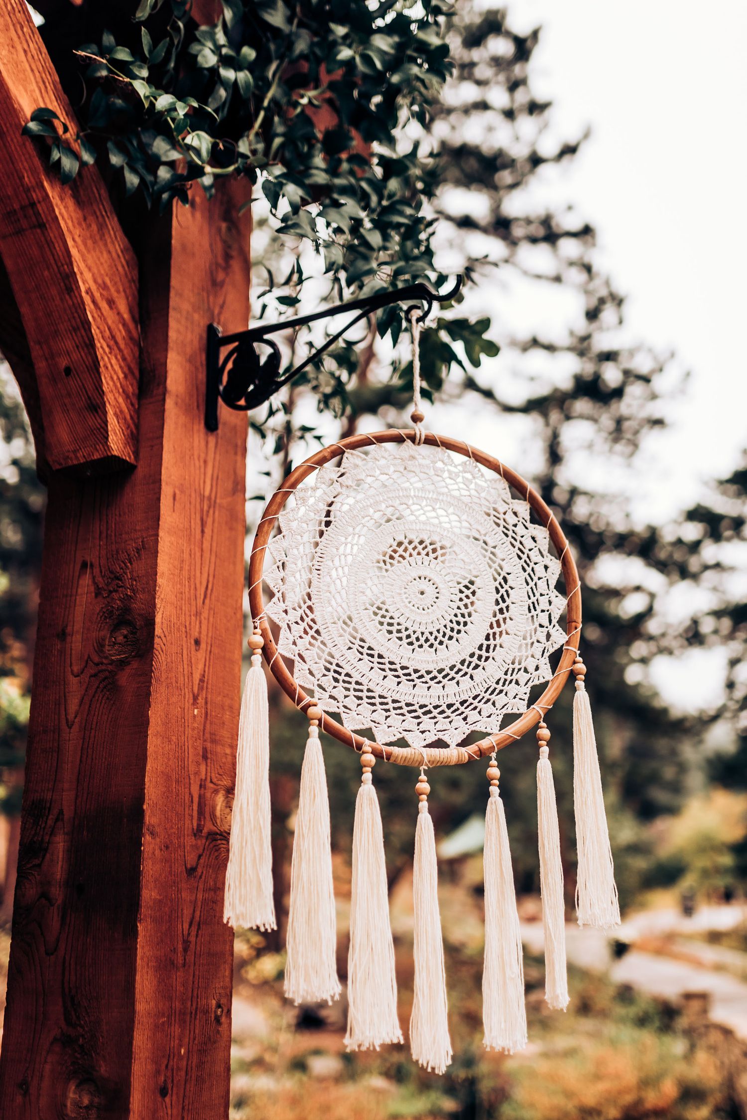 Boho dreamcatcher with white tassels hanging from wooden post against forest backdrop.