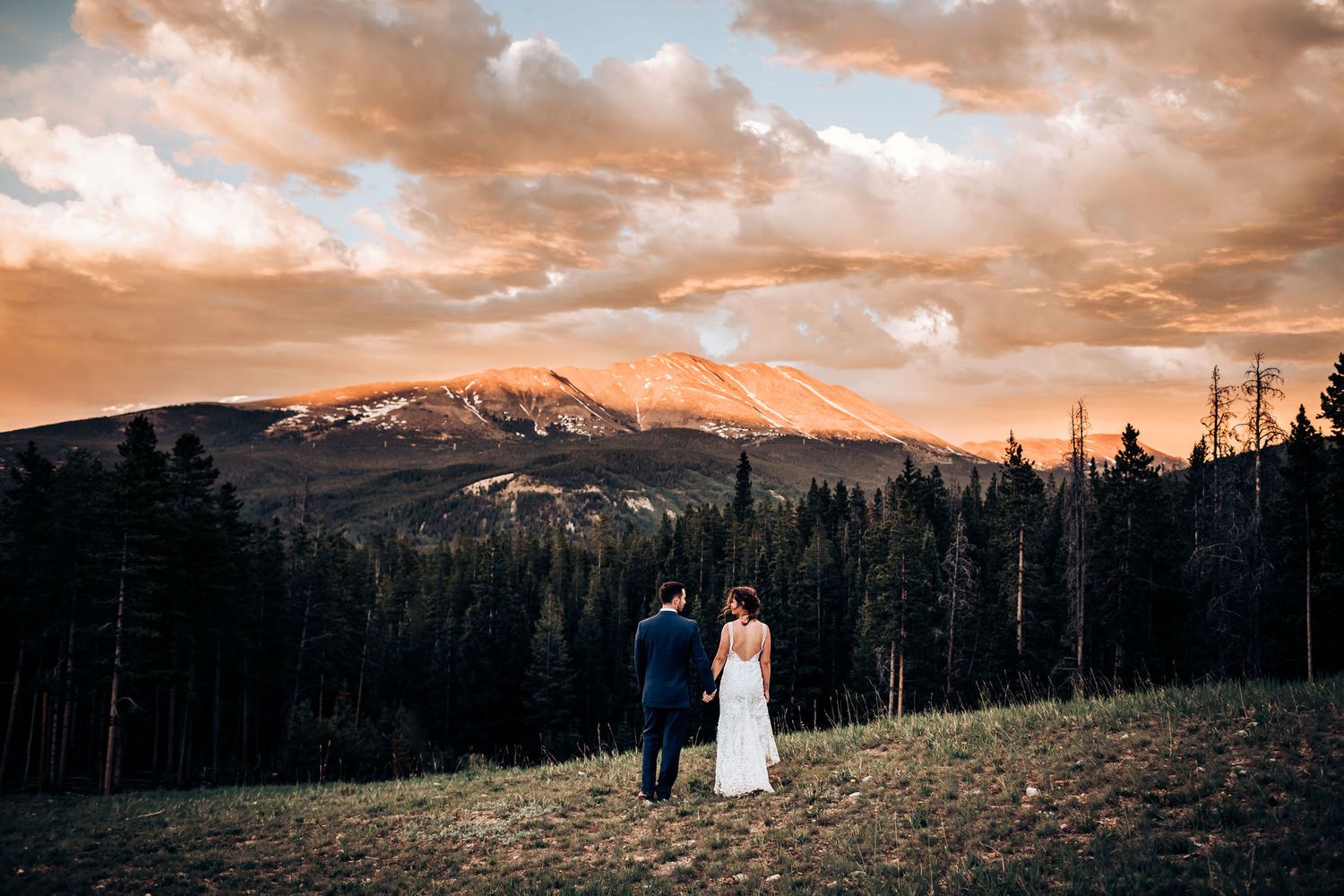 A wedding couple silhouetted against dramatic sunset sky and mountain landscape.