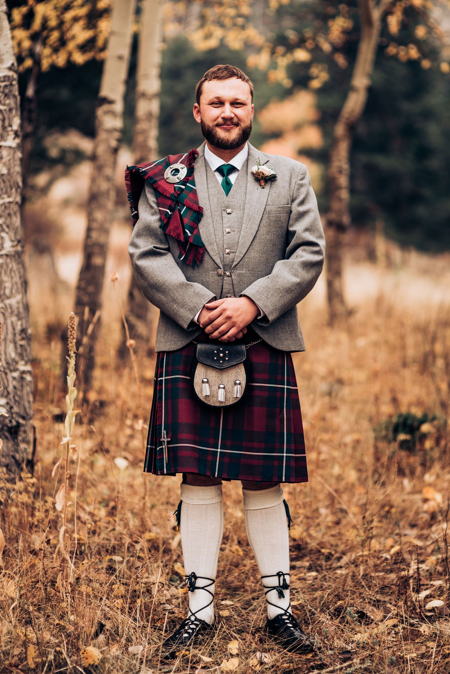Groom in traditional Scottish attire with grey suit jacket and tartan kilt standing in autumn setting.