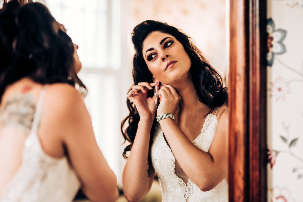 A bride in a white dress putting on earrings while looking in a mirror.