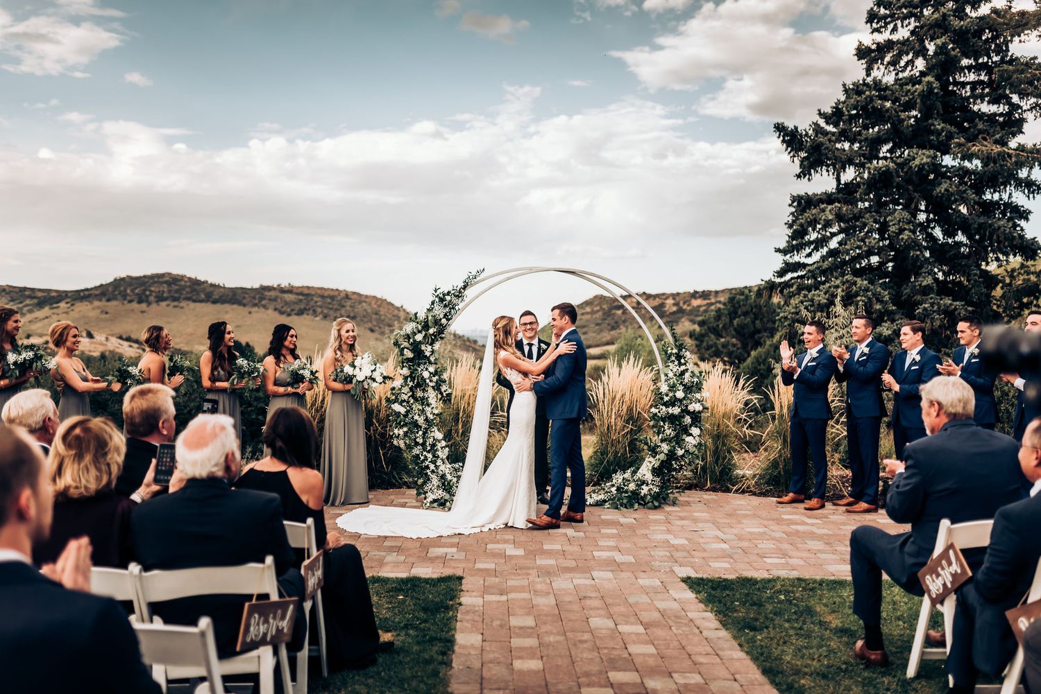 Wide angle view of mountain wedding ceremony with guests seated facing decorated circular arch.