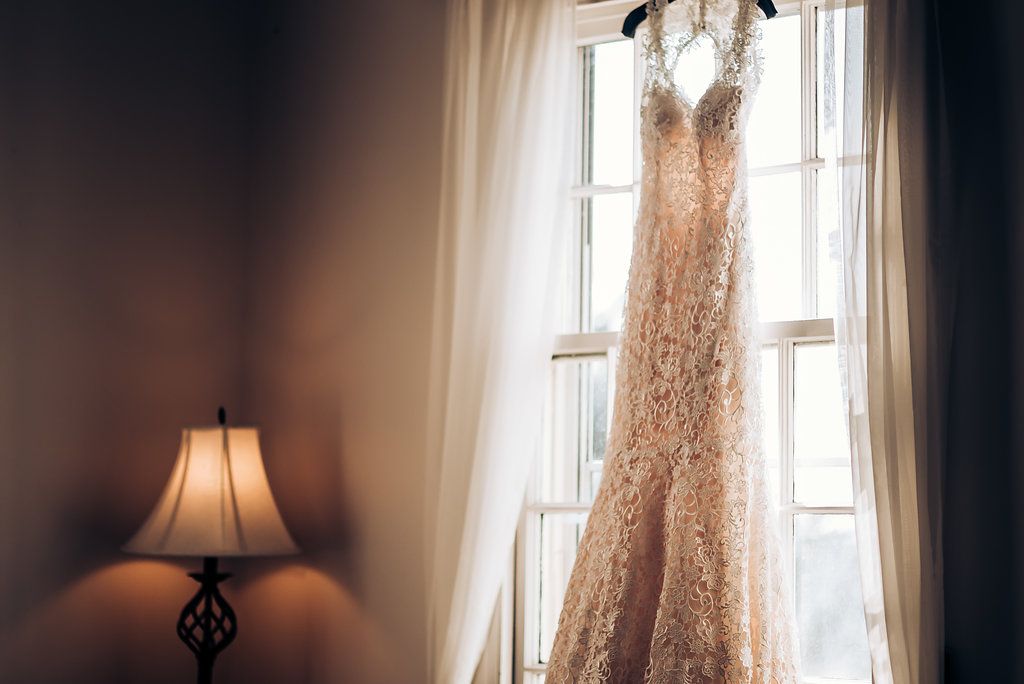 Elegant lace wedding dress hanging in natural window light against white curtains.