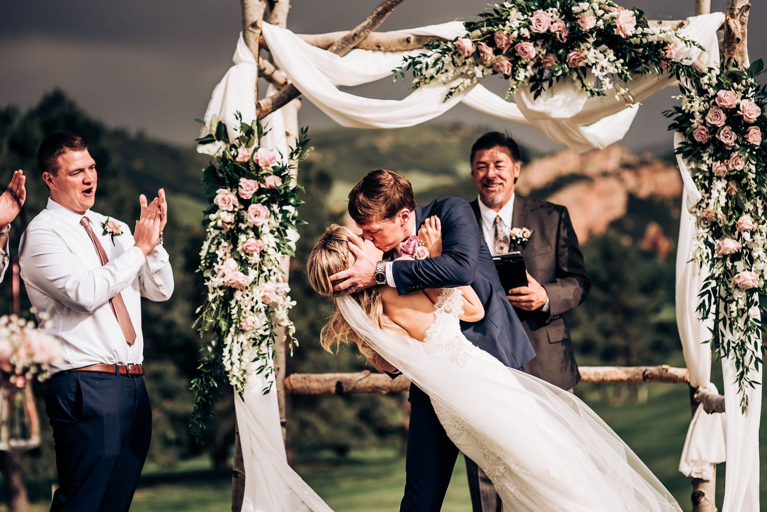 Romantic outdoor wedding ceremony under a floral arch decorated with white fabric and pink roses in a mountain setting.