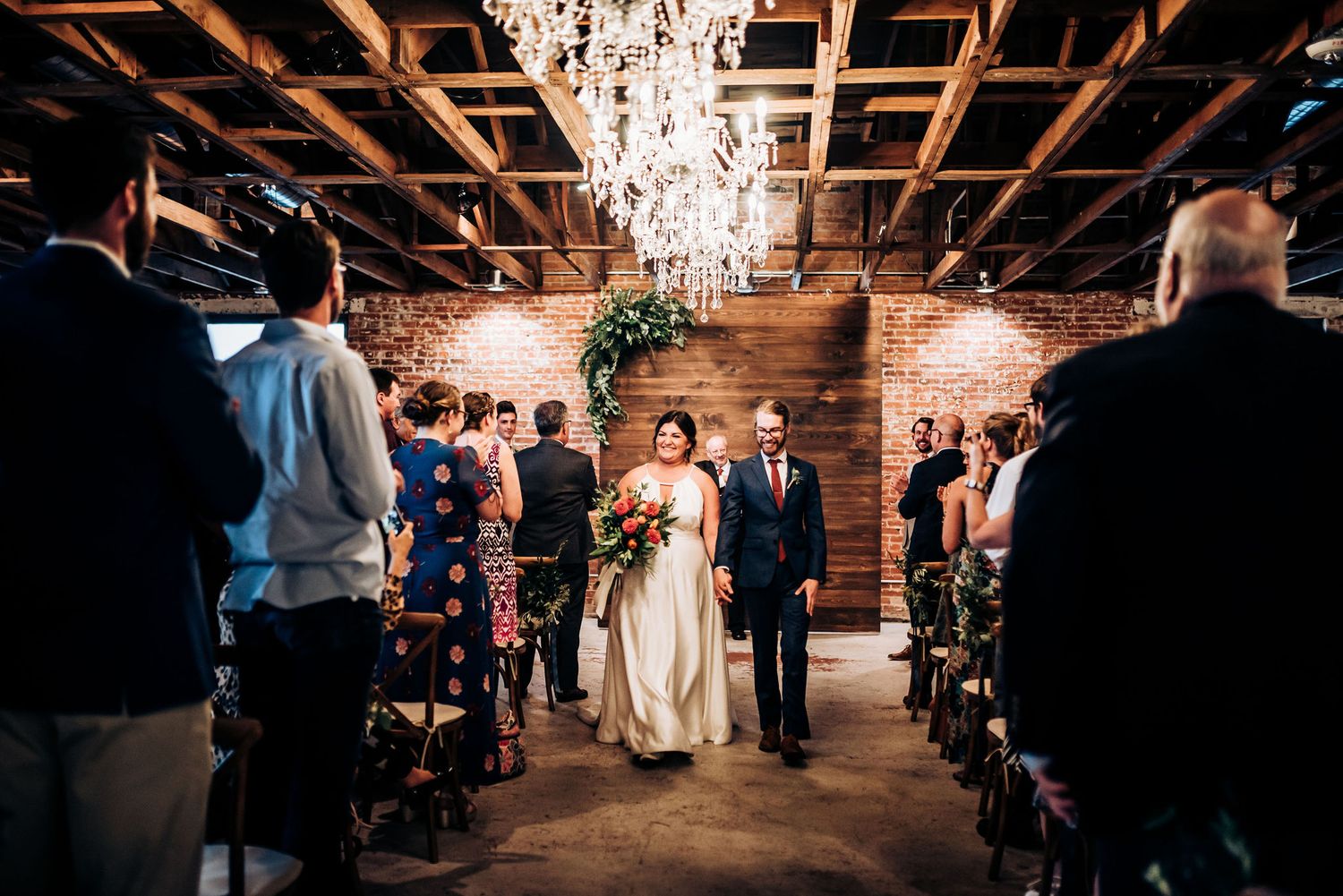 Wedding processional in industrial venue with exposed beams and crystal chandelier overhead.