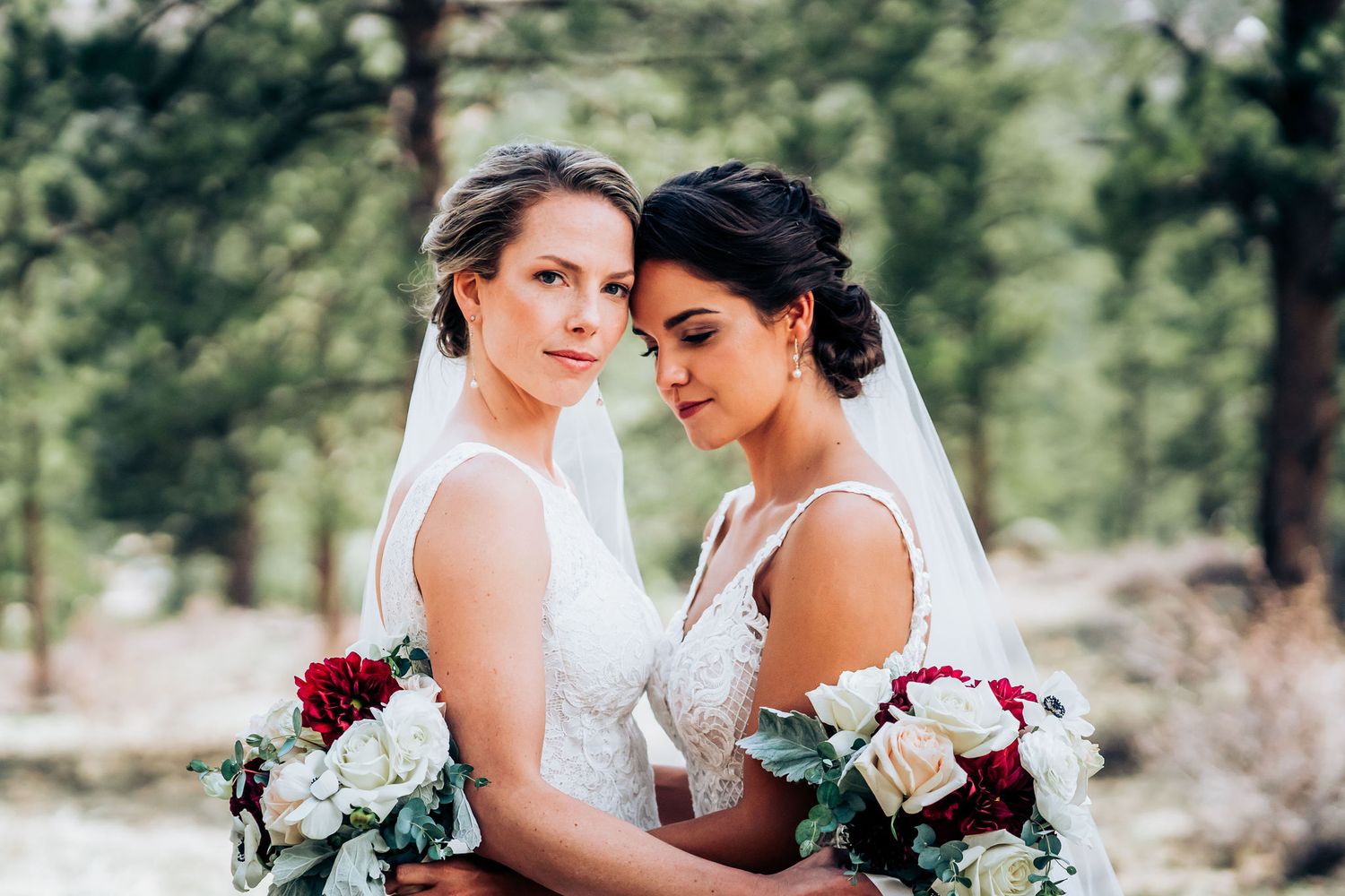 Close-up portrait of two brides with elegant bouquets against forest background.