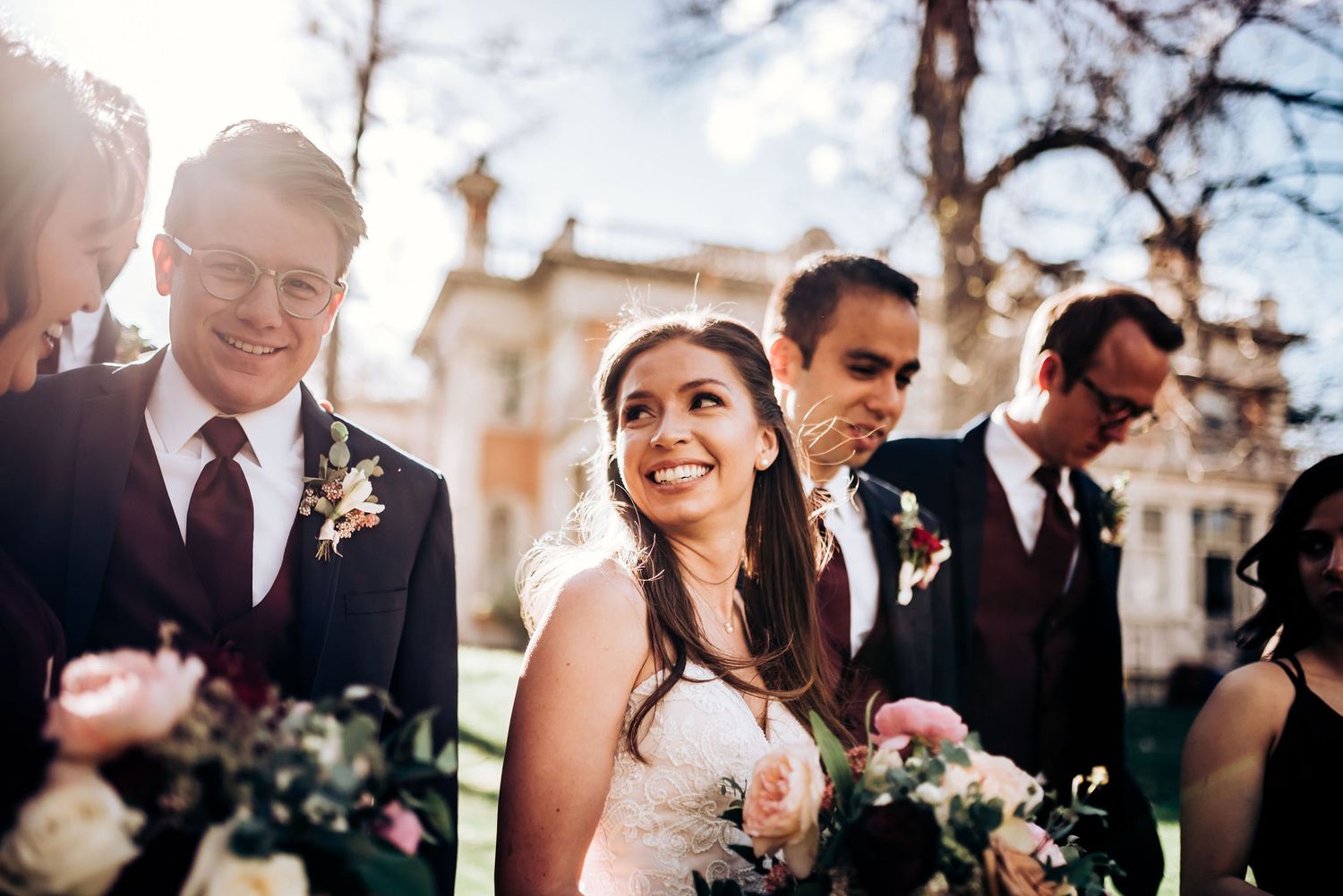 Wedding party members share joyful laughter together outdoors with sunlight streaming through trees.