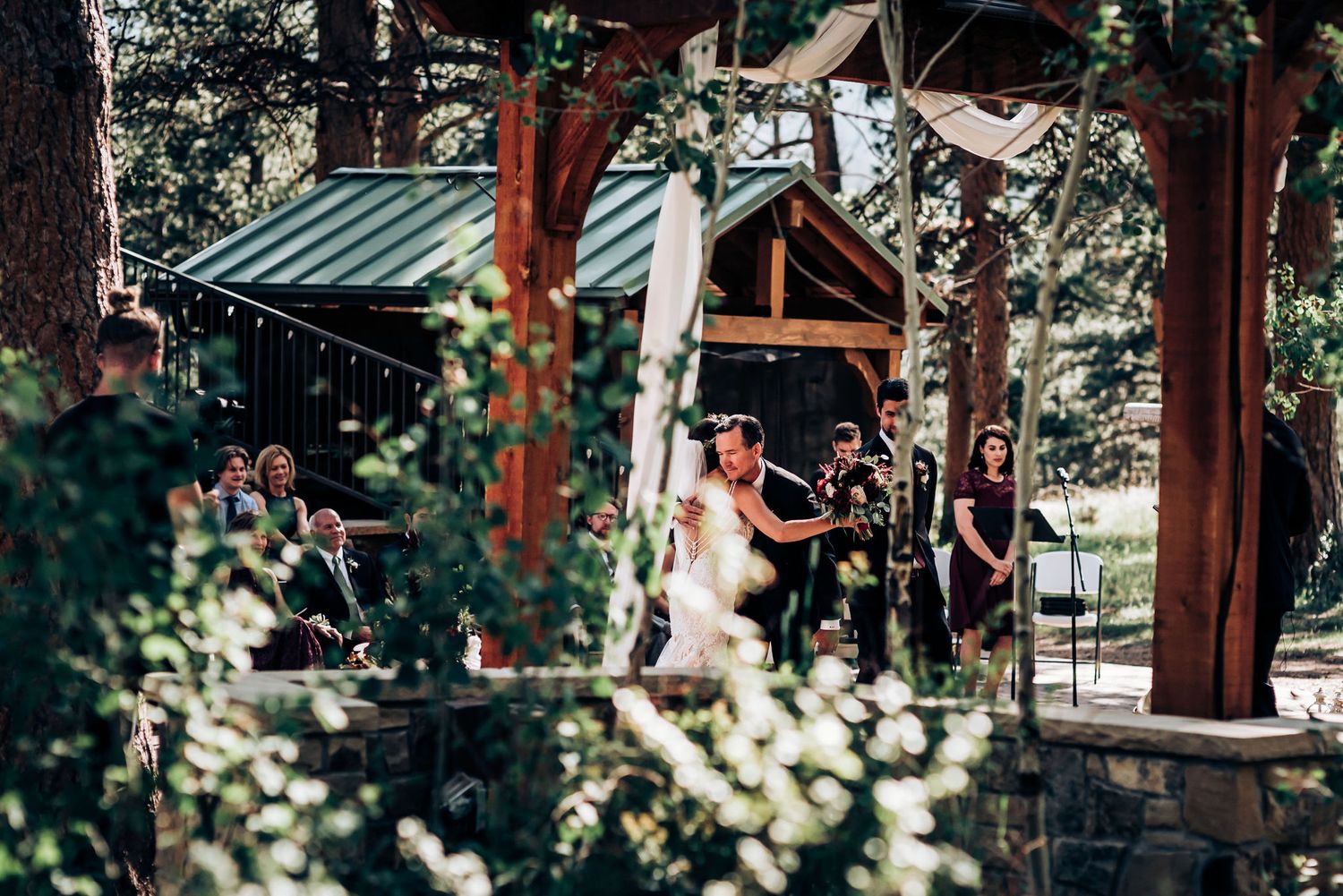 An outdoor wedding ceremony under a wooden pavilion surrounded by forest scenery.