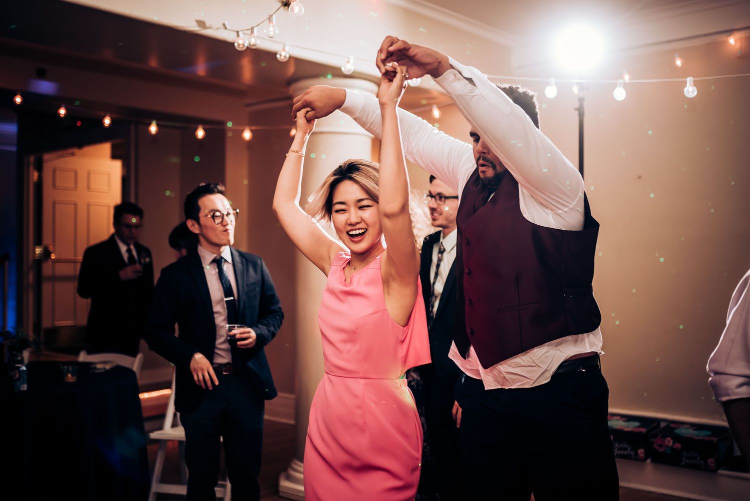 Guests dancing energetically at a wedding reception with string lights in the background.