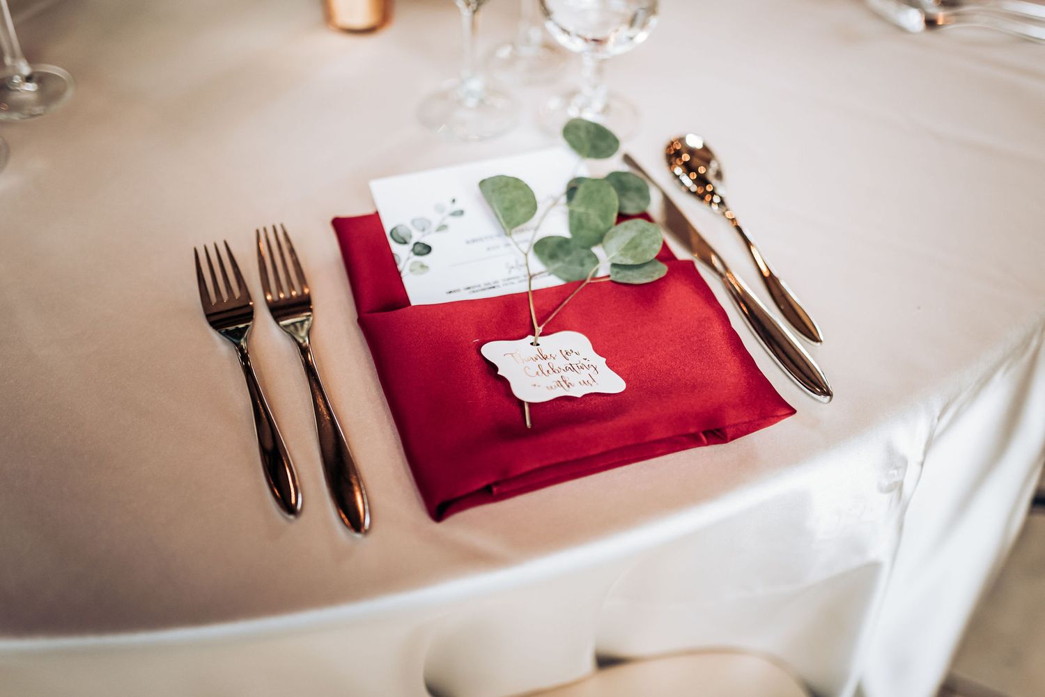 Elegant place setting with red napkin, silverware and eucalyptus on white tablecloth.