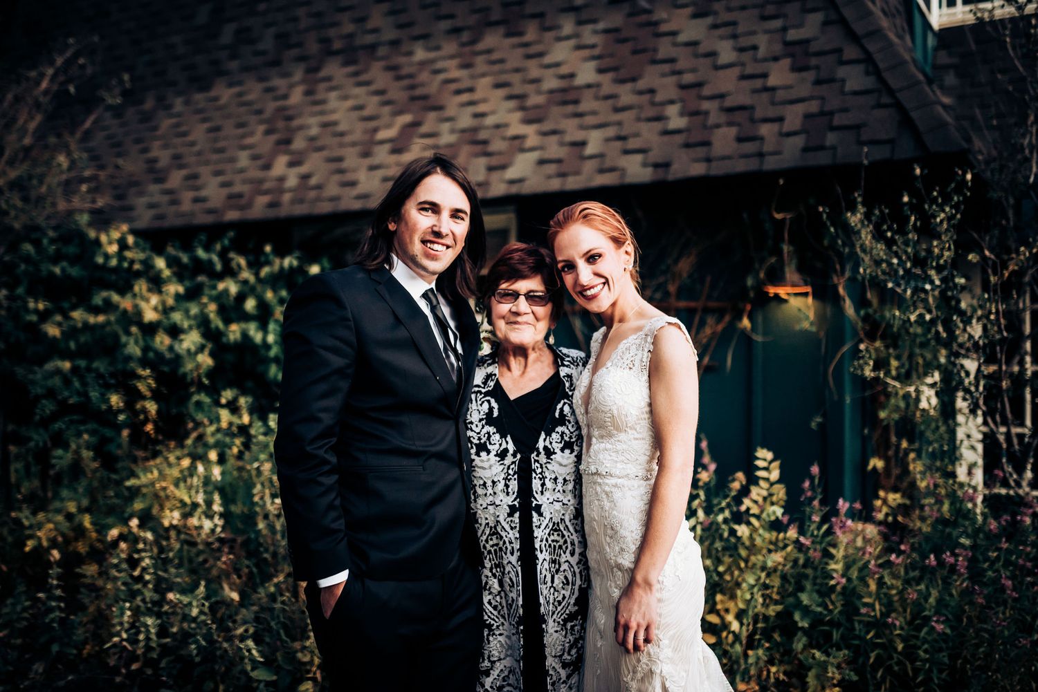 A wedding party poses together in front of a house with a garden backdrop.