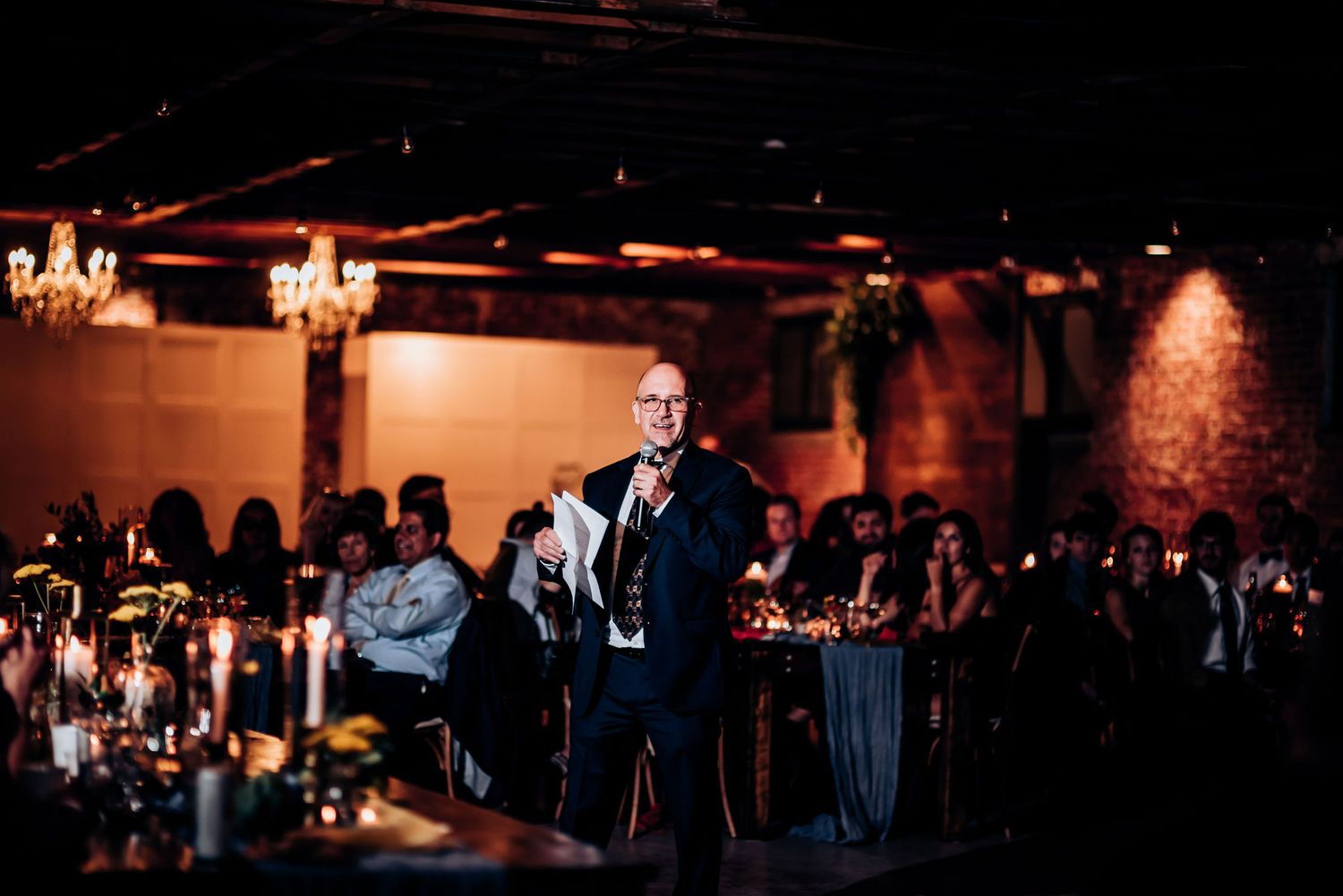 A speaker addresses guests at an elegantly lit indoor venue with chandeliers and brick walls.