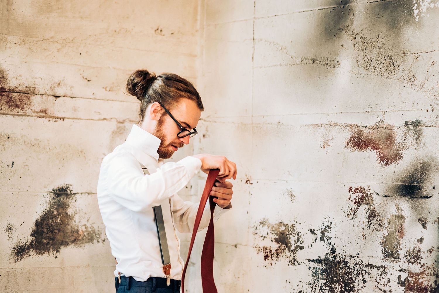 Someone adjusts a burgundy tie while standing against a distressed wall in a white dress shirt.