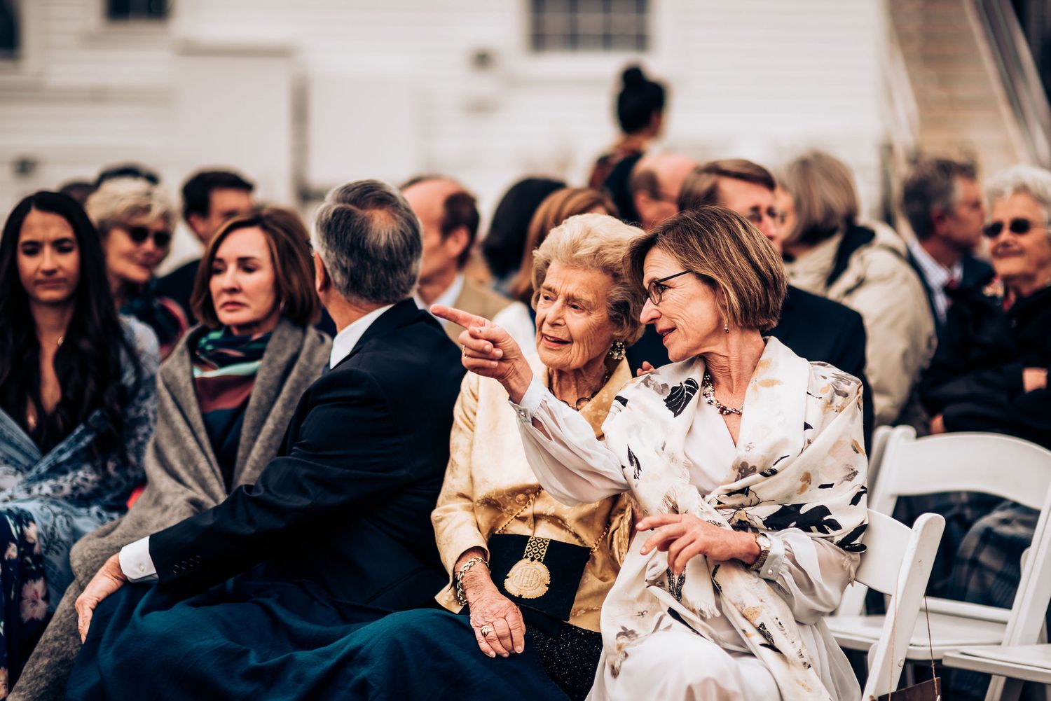 Wedding guests seated in white chairs outdoors watching ceremony at elegant venue.
