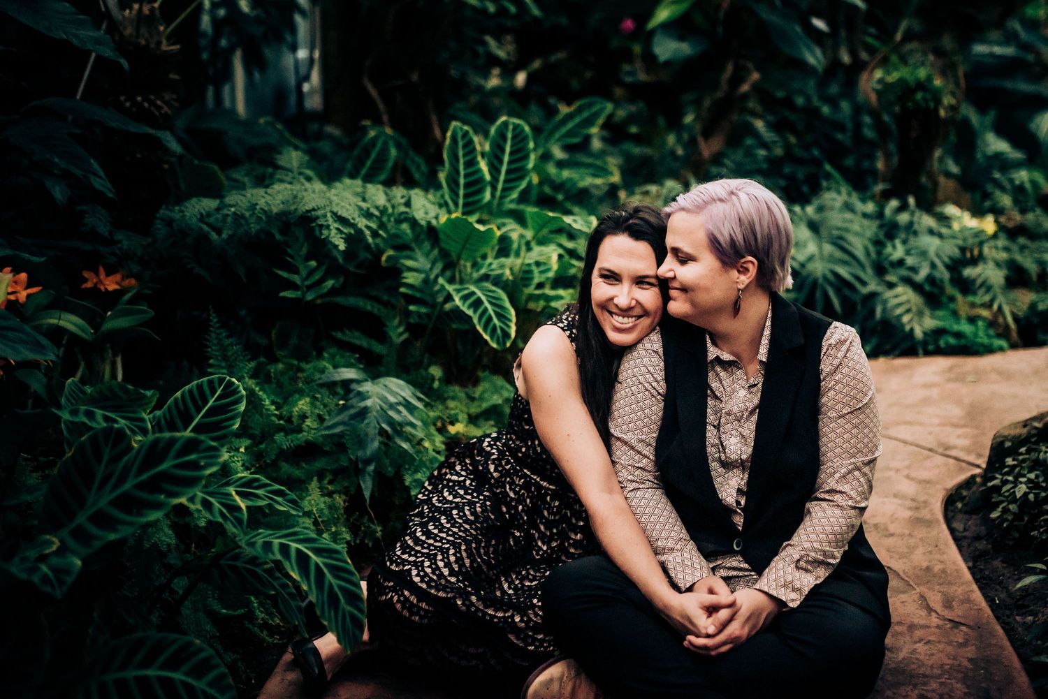 A couple sits close together on a garden path surrounded by tropical plants.