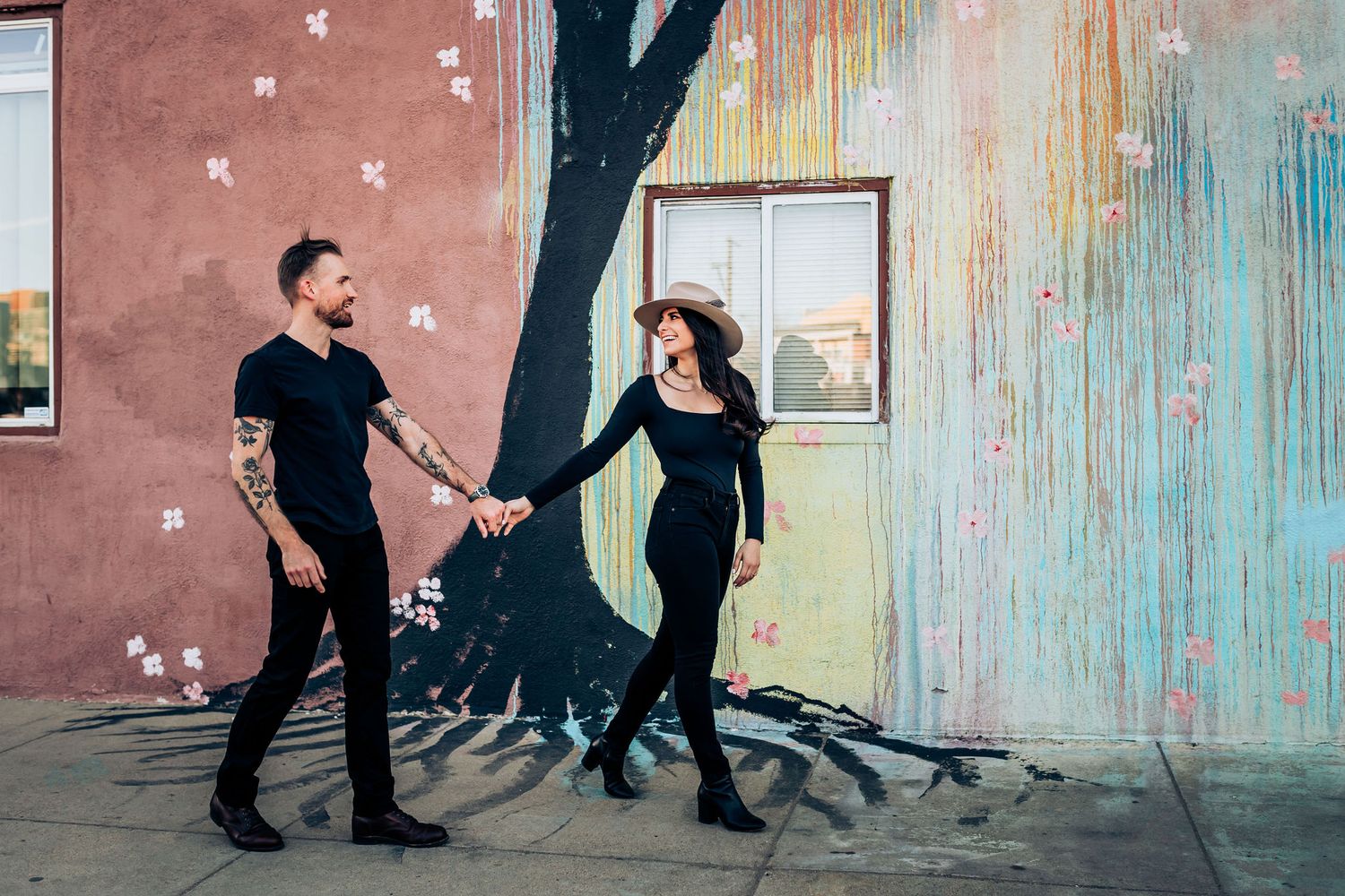 Couple dances playfully against pastel-colored wall with artistic designs.