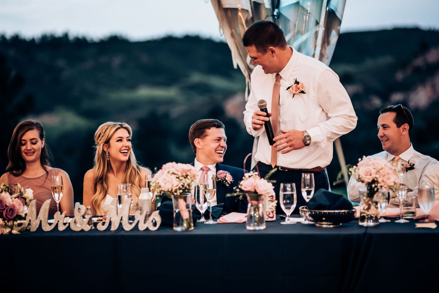 A wedding reception speech being given at an outdoor venue with guests seated at a long table during sunset.
