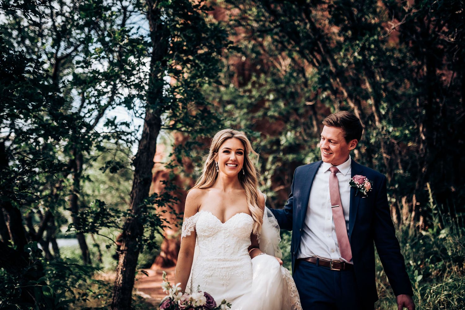 Natural outdoor wedding portrait with couple walking through wooded area carrying pink bouquet.