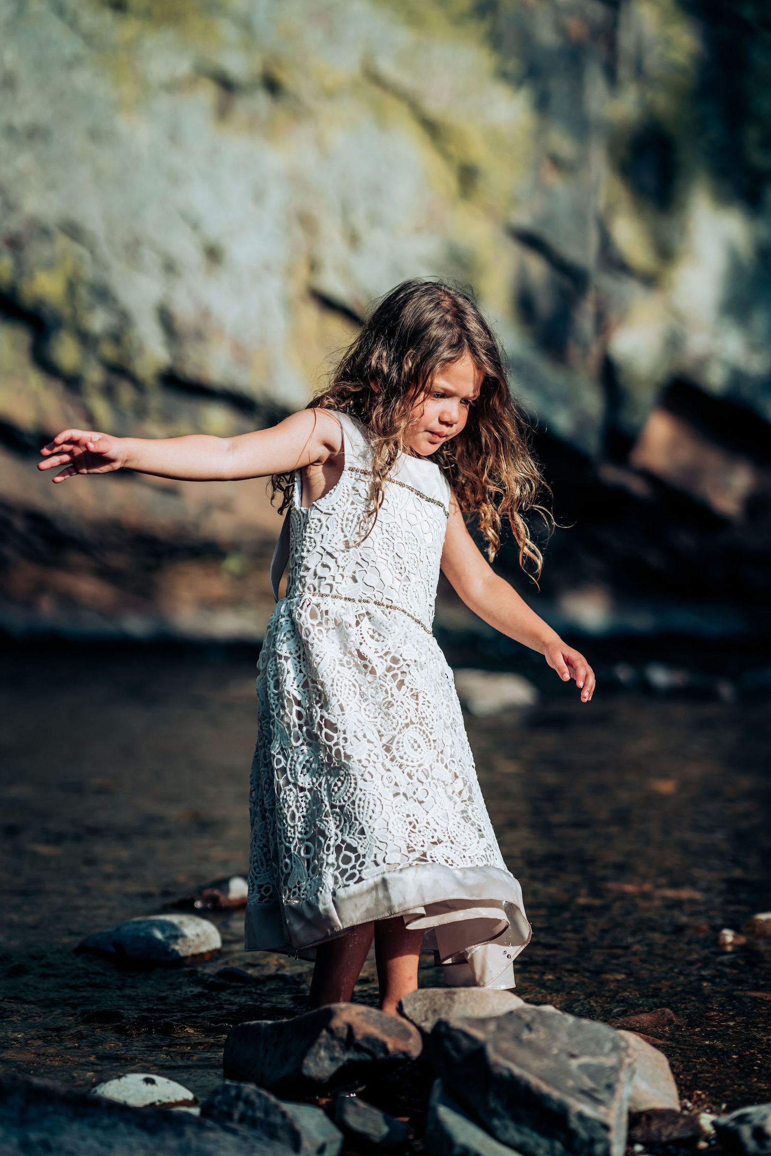 Young girl in white lace dress dances on rocks by water with cliffs in background.