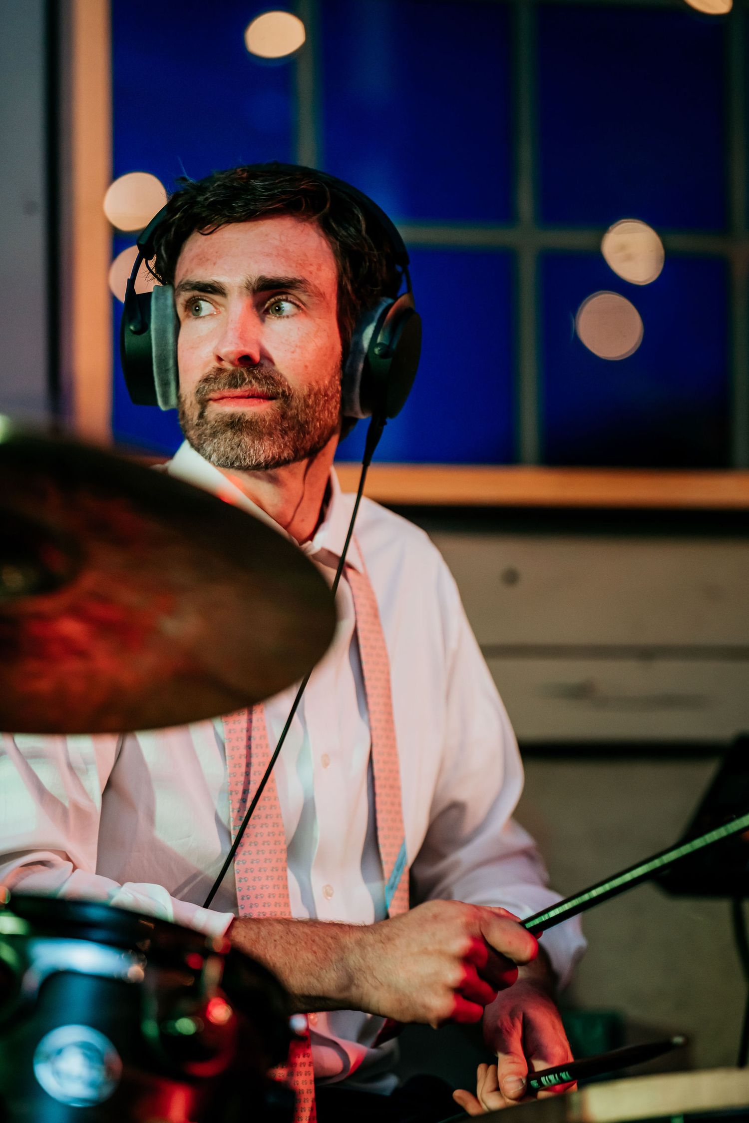 Drummer wearing headphones and dress shirt performs on stage with dramatic lighting.